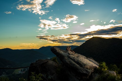 A couple sits on a large rock at sunset, overlooking a valley with mountains and colorful clouds in the sky.