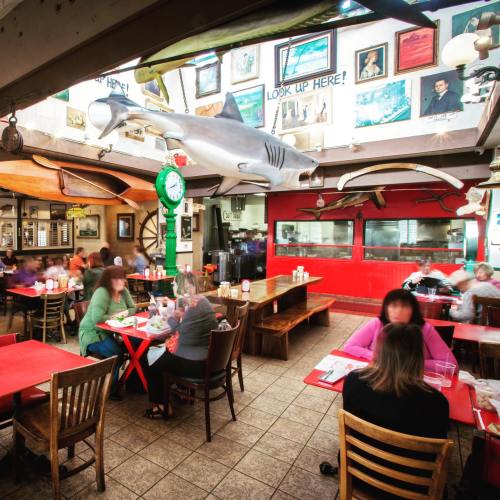 A bustling restaurant interior with red tables, a large shark model hanging, and patrons enjoying their meals.