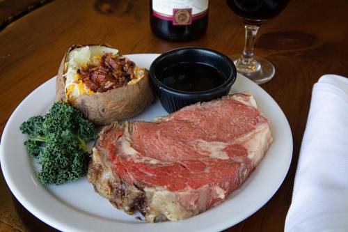 A plate with a juicy prime rib, baked potato with toppings, steamed kale, and a glass of red wine in the background.