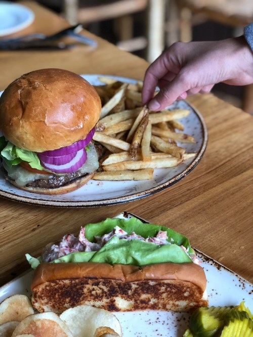 A hand reaches for fries next to a burger and a lobster roll on a wooden table.