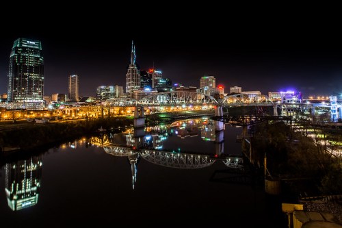 City skyline at night with illuminated buildings and a bridge reflected in the water below.