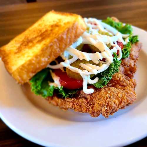 A crispy fried chicken sandwich with lettuce, tomato, pickles, and mayonnaise, served with toasted bread on a plate.