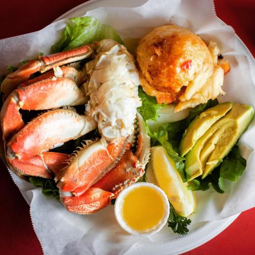 A plate featuring crab claws, a seafood cake, avocado slices, lettuce, lemon, and a small cup of sauce.