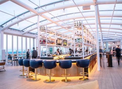 A modern bar with a sleek design, featuring high stools, a well-stocked shelf, and large windows showcasing a view.