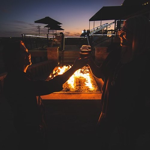 Two people toast with drinks in front of a fire pit at sunset, creating a warm and inviting atmosphere.