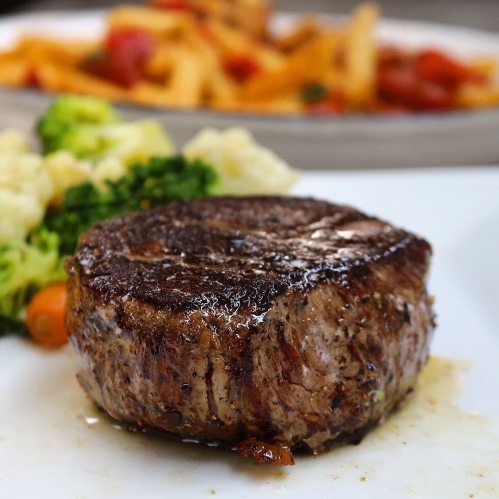 A perfectly cooked filet mignon on a plate, accompanied by colorful vegetables and a blurred pasta dish in the background.