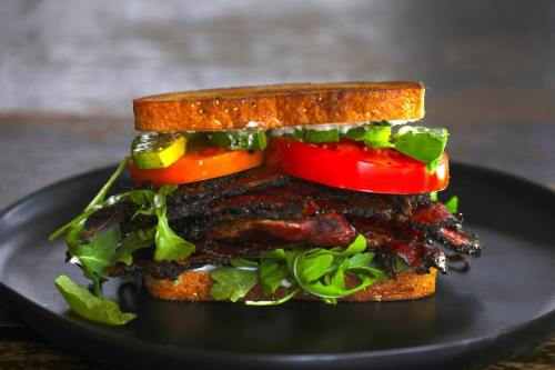 A hearty sandwich with layers of grilled meat, fresh tomatoes, pickles, and greens, served on whole grain bread.