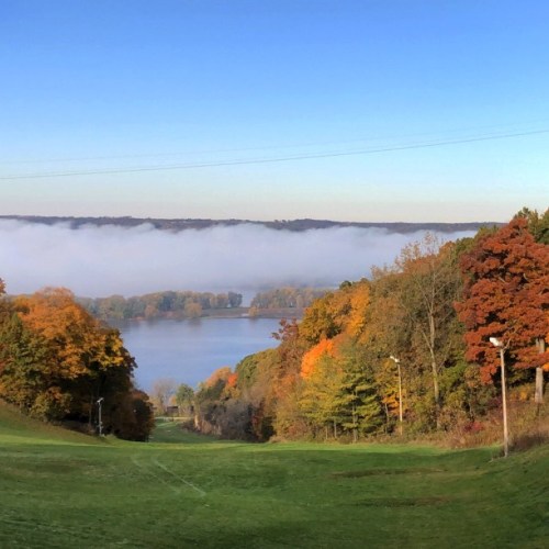 A scenic view of a river surrounded by colorful autumn trees and fog, with a clear blue sky above.