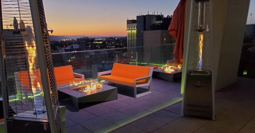 A rooftop lounge with orange seating and fire features, overlooking a city skyline at sunset.