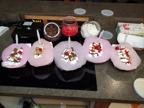 Five dessert bowls topped with cotton candy, whipped cream, chocolate pieces, cherries, and sprinkles on a countertop.