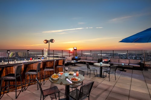 Rooftop bar with tables, drinks, and a sunset view over the city skyline.