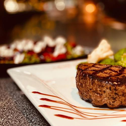 A perfectly grilled steak on a white plate, garnished with sauce, with a colorful salad in the background.