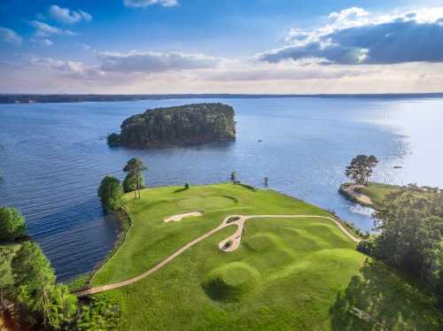 Aerial view of a golf course by a lake, featuring green fairways and an island in the background under a blue sky.