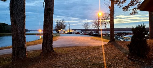 A serene lakeside scene at dusk, with parked cars and a building illuminated by warm lights among trees.