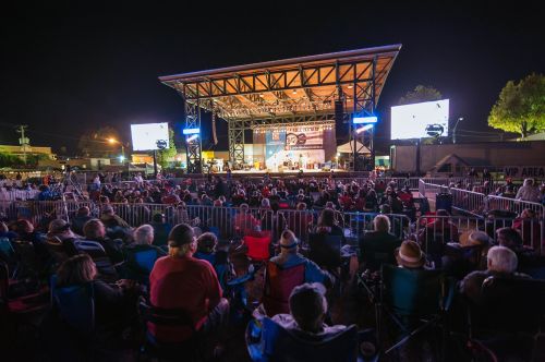 A large crowd enjoys a nighttime concert at an outdoor stage, with bright lights and screens displaying the performance.