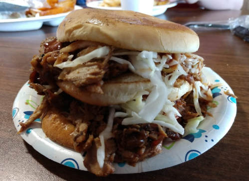 A stacked pulled pork sandwich with coleslaw on a paper plate, set on a wooden table.
