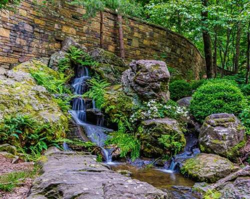 A serene waterfall cascades over rocks, surrounded by lush greenery and a stone wall in a tranquil garden setting.
