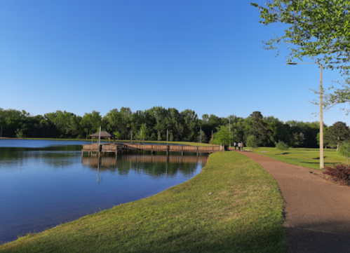 A serene park scene with a calm lake, a wooden dock, and a walking path lined with trees under a clear blue sky.