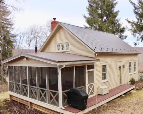 A charming beige house with a metal roof and a screened porch, surrounded by trees and a grassy area.