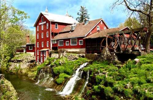 A red historic mill beside a stream with small waterfalls, surrounded by greenery and trees under a blue sky.