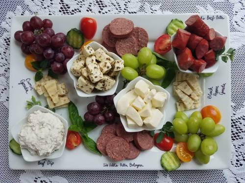 A colorful cheese and meat platter featuring grapes, tomatoes, cucumbers, and various dips in small bowls.