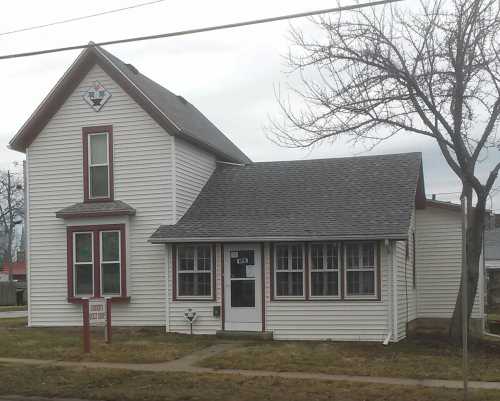 A two-story house with a peaked roof, white siding, red trim, and a sign in front, set in a grassy area.