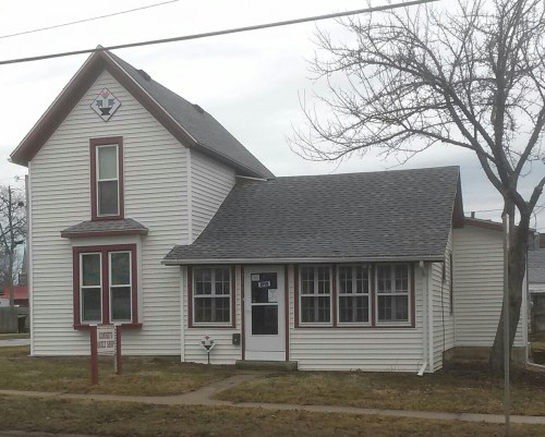 A two-story house with a peaked roof, white siding, red trim, and a sign in front, set in a grassy area.