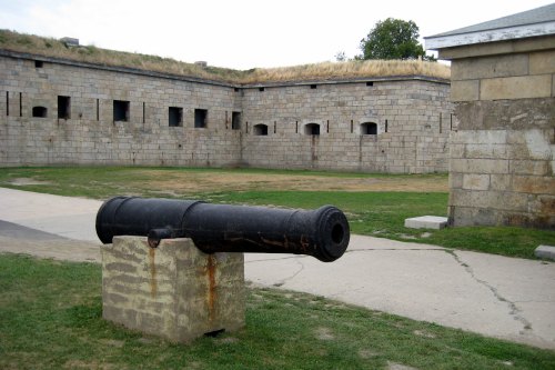 A historic cannon on a stone pedestal in a grassy courtyard surrounded by stone fort walls.