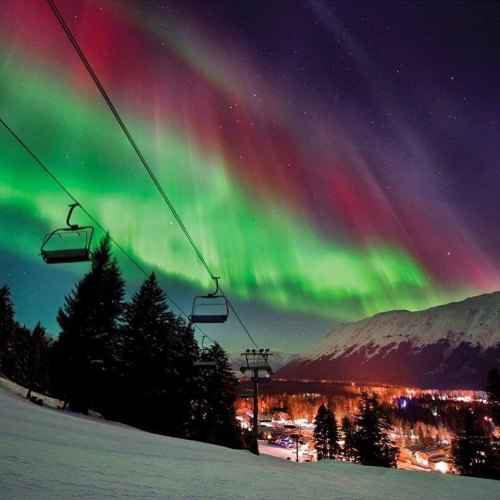 A stunning display of the Northern Lights over a snowy landscape, with ski lift cables and a mountain in the background.