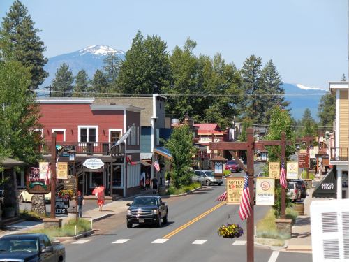 A charming small town street lined with shops, trees, and mountains in the background on a sunny day.