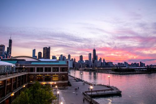 Sunset over a city skyline with colorful clouds, reflecting on a river, and a waterfront promenade below.