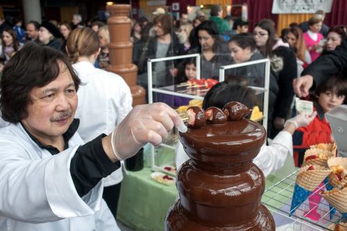 A chocolatier adds treats to a chocolate fountain at a busy event, with a crowd of people in the background.