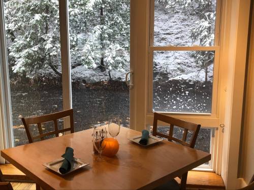 A cozy dining table set for two, with a pumpkin centerpiece, overlooking a snowy landscape through large windows.