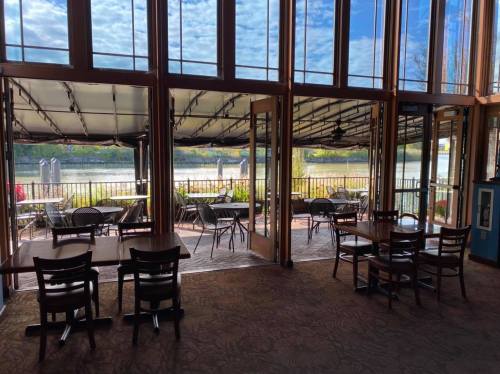 Interior view of a restaurant with large windows overlooking a patio and river, featuring empty tables and chairs.
