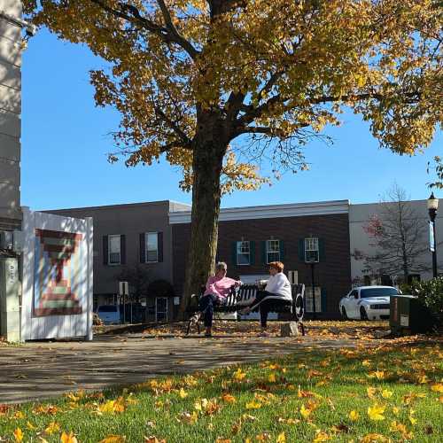 Two people sit on a bench under a large tree with autumn leaves, surrounded by a quaint town setting.