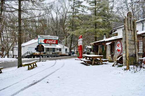 A snowy scene of a general store with vintage signage, surrounded by trees and rustic buildings.