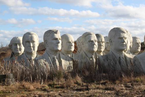 A row of large, unfinished stone heads of various historical figures, surrounded by tall grass under a cloudy sky.