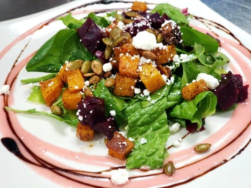 A vibrant salad with mixed greens, roasted butternut squash, beets, pumpkin seeds, and feta cheese drizzled with dressing.