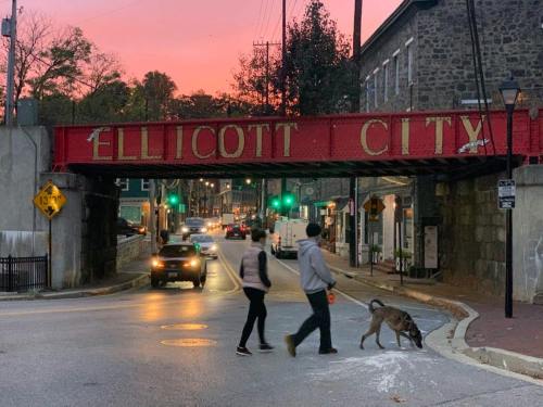 Two people and a dog cross the street under a red bridge sign reading "Ellicott City" at sunset.