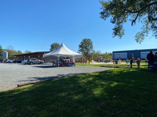 A sunny outdoor scene with a tent, cars parked, and people gathered in a grassy area near a building.