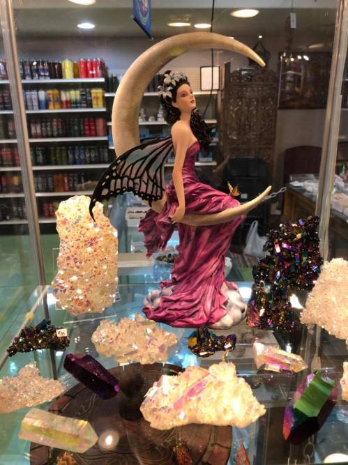 A fairy figurine in a purple dress sits on a crescent moon, surrounded by colorful crystals and stones in a display case.