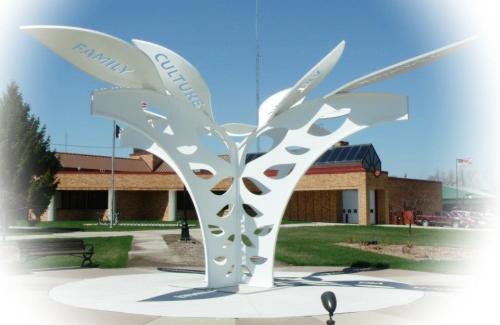 A white sculpture resembling a tree with leaves labeled "Family" and "Cultures," set in a grassy area near a building.