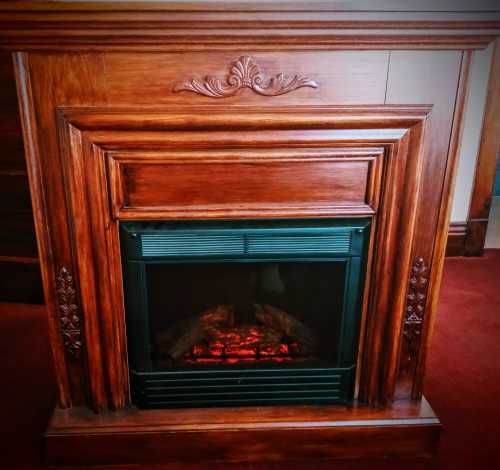 A wooden fireplace with ornate detailing and glowing logs inside, set against a red carpet background.