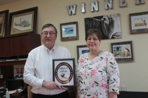 A man and woman stand together, holding a plaque that reads "Keep It Local," in an office setting.