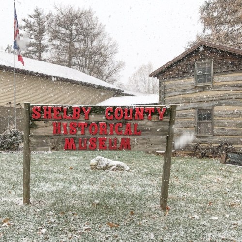 A snowy scene featuring a sign for the Shelby County Historical Museum in front of a rustic building.