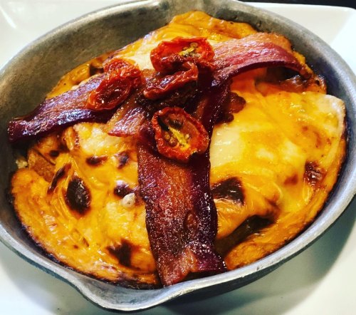 A baked dish with melted cheese, crispy bacon, and sun-dried tomatoes in a small metal skillet.