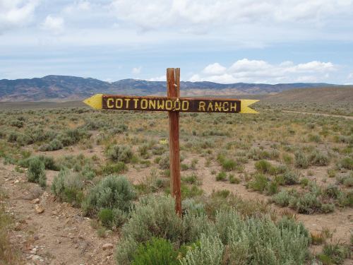 A wooden signpost pointing to "Cottonwood Ranch" in a grassy, arid landscape with mountains in the background.