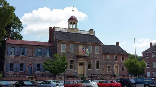 Historic building with a dome, flags, and brick facade, surrounded by trees and parked cars under a blue sky.