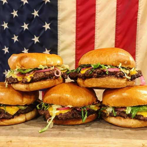 A stack of delicious burgers with cheese, lettuce, and onions in front of an American flag backdrop.