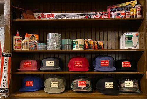 A shelf displaying various hats with labels, glue, tape, and other tools, creating a vintage store atmosphere.